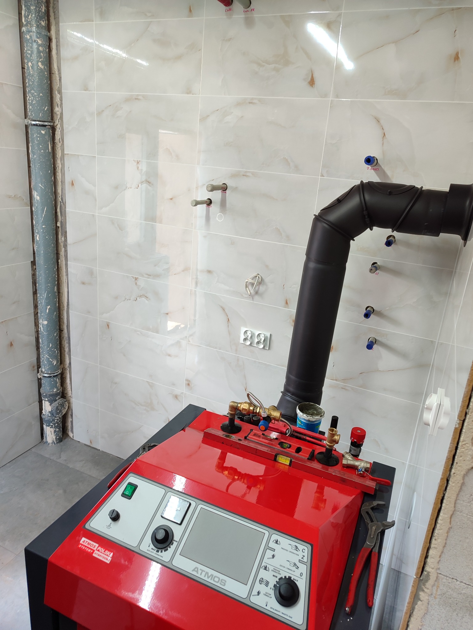 Czerwony piec Atmos z czarnym kominem w trakcie instalacji, widoczne narzędzia hydrauliczne, rury doprowadzające oraz wykończona ściana z jasnych płytek imitujących marmur.