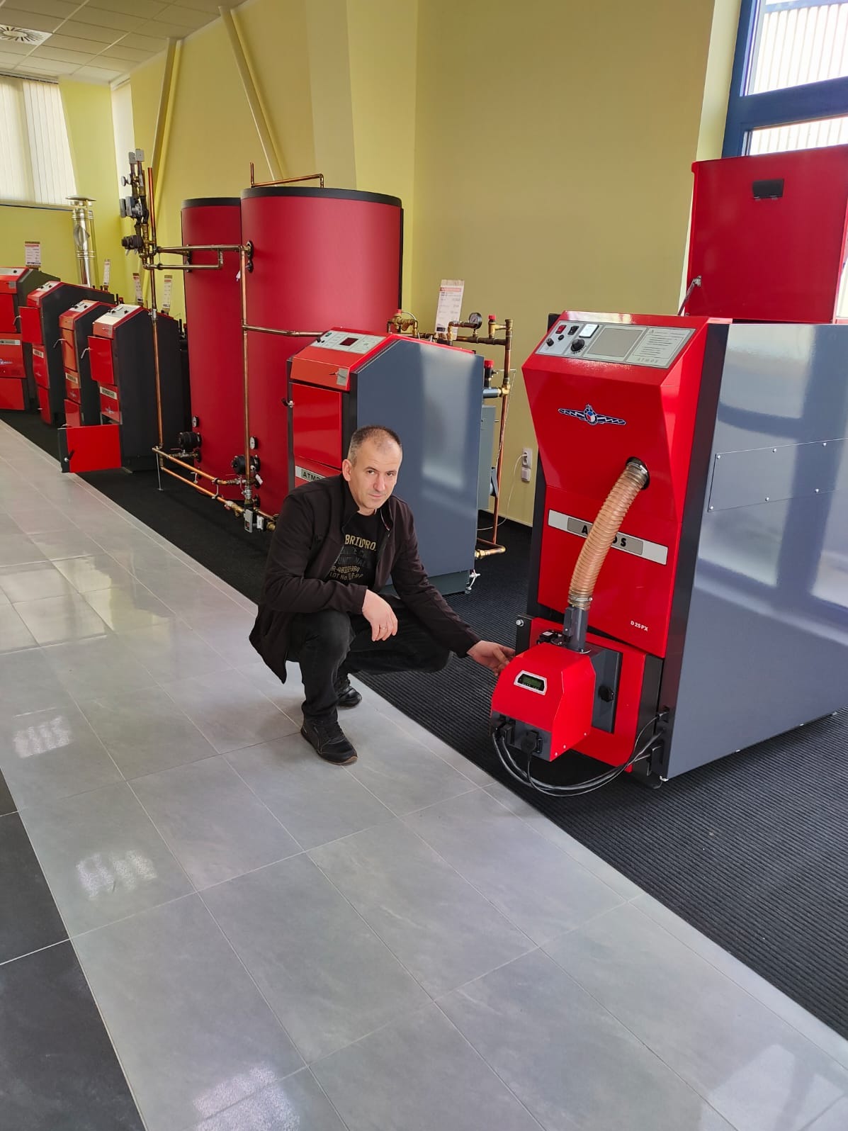 Mężczyzna kuca obok kotła na paliwo stałe Atmos D 25 PX w showroomie, prezentując jego panel sterowania. W tle rzędy innych kotłów i zbiorników akumulacyjnych w kolorach czerwonym i szarym.