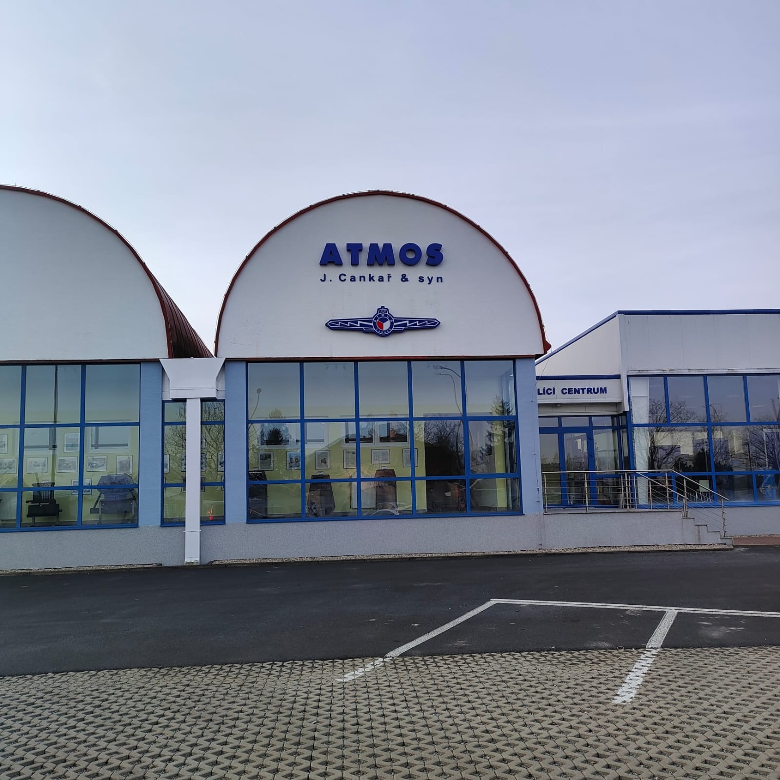 Budynek firmy Atmos J. Cankař & syn z niebieskim logo i charakterystycznym znakiem graficznym nad wejściem, widok z zewnątrz na parking i kostkę brukową.