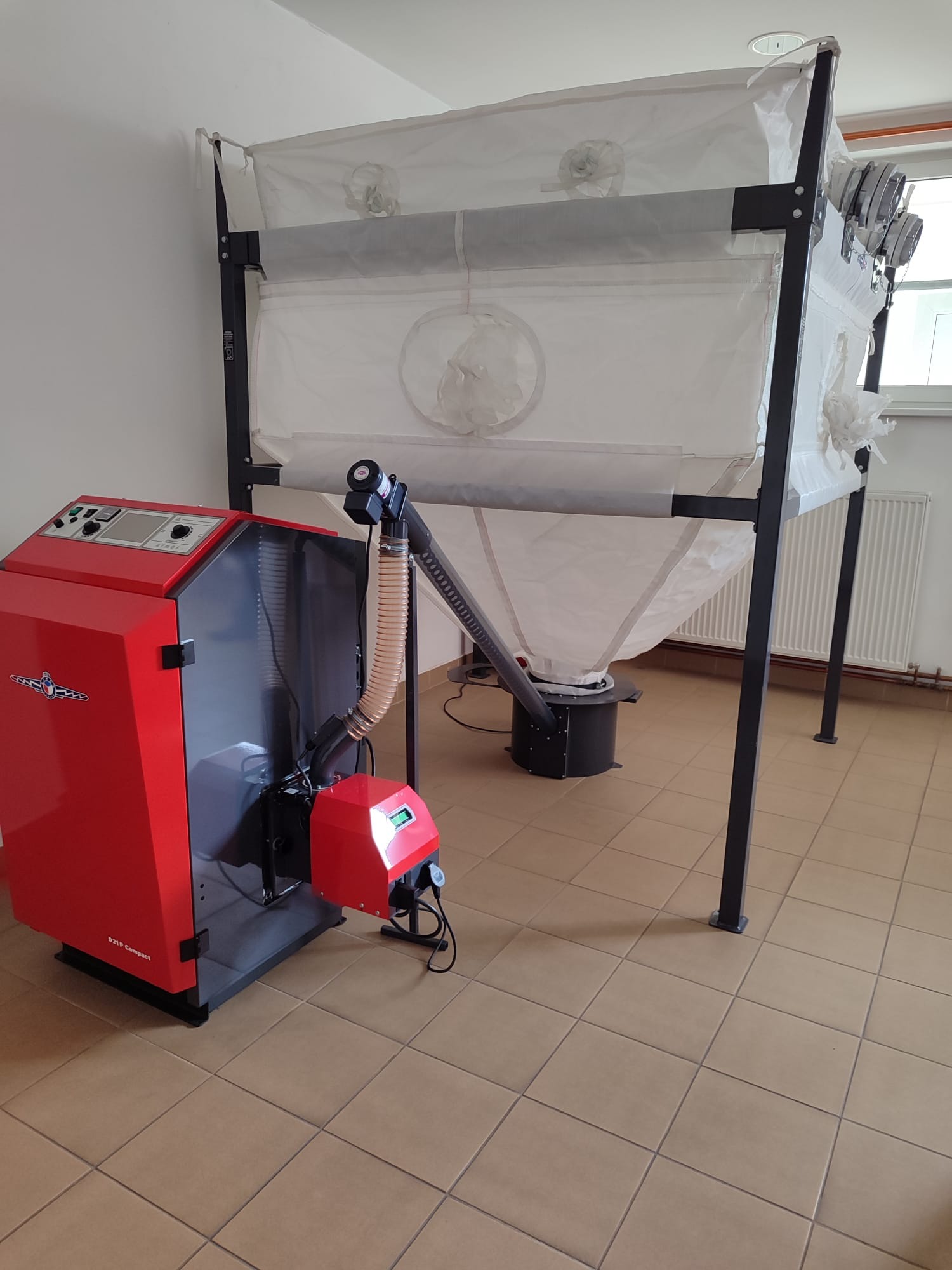 Czerwono-szary piec na paliwo stałe z podajnikiem i zasobnikiem na pellet, widoczne elementy sterujące i podłączenie do podajnika, na brązowej podłodze z płytek.