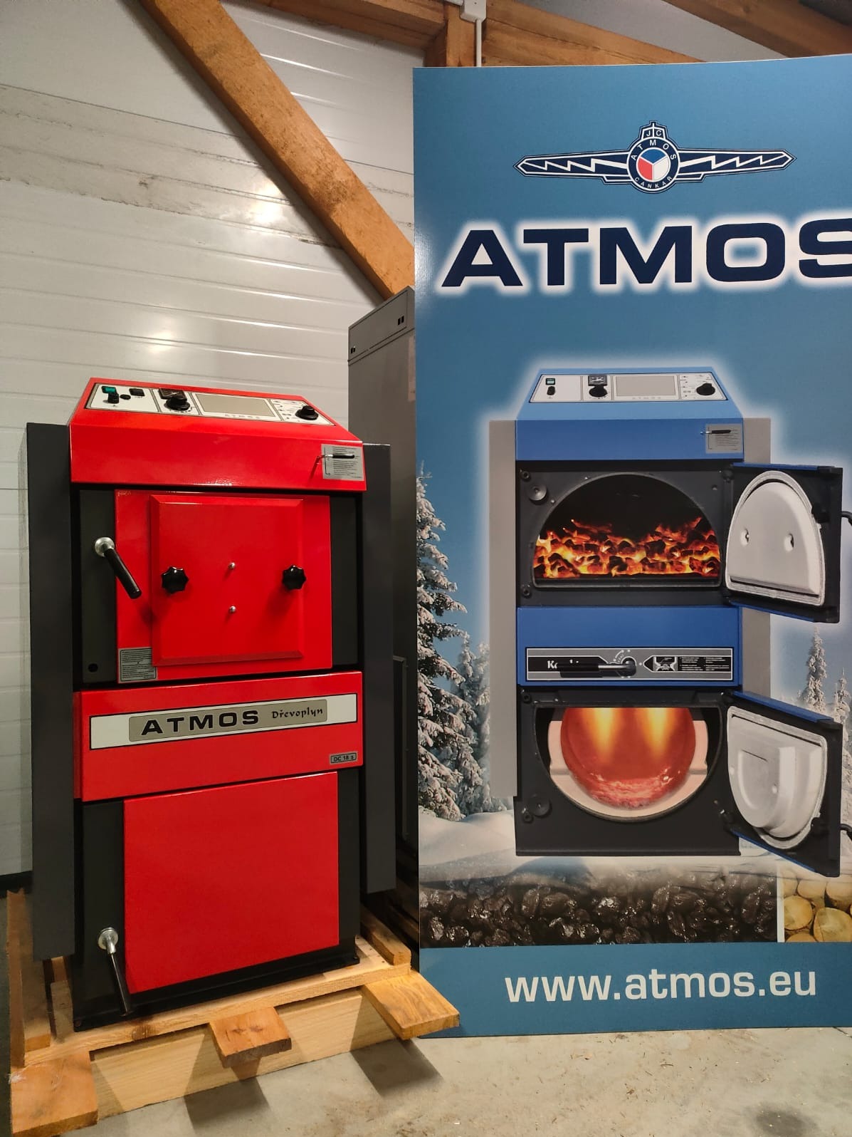 Czerwony piec Atmos Dřevoplyn na palecie, obok reklama z niebieskim piecem Atmos, na tle biała ściana z drewnianą belką.