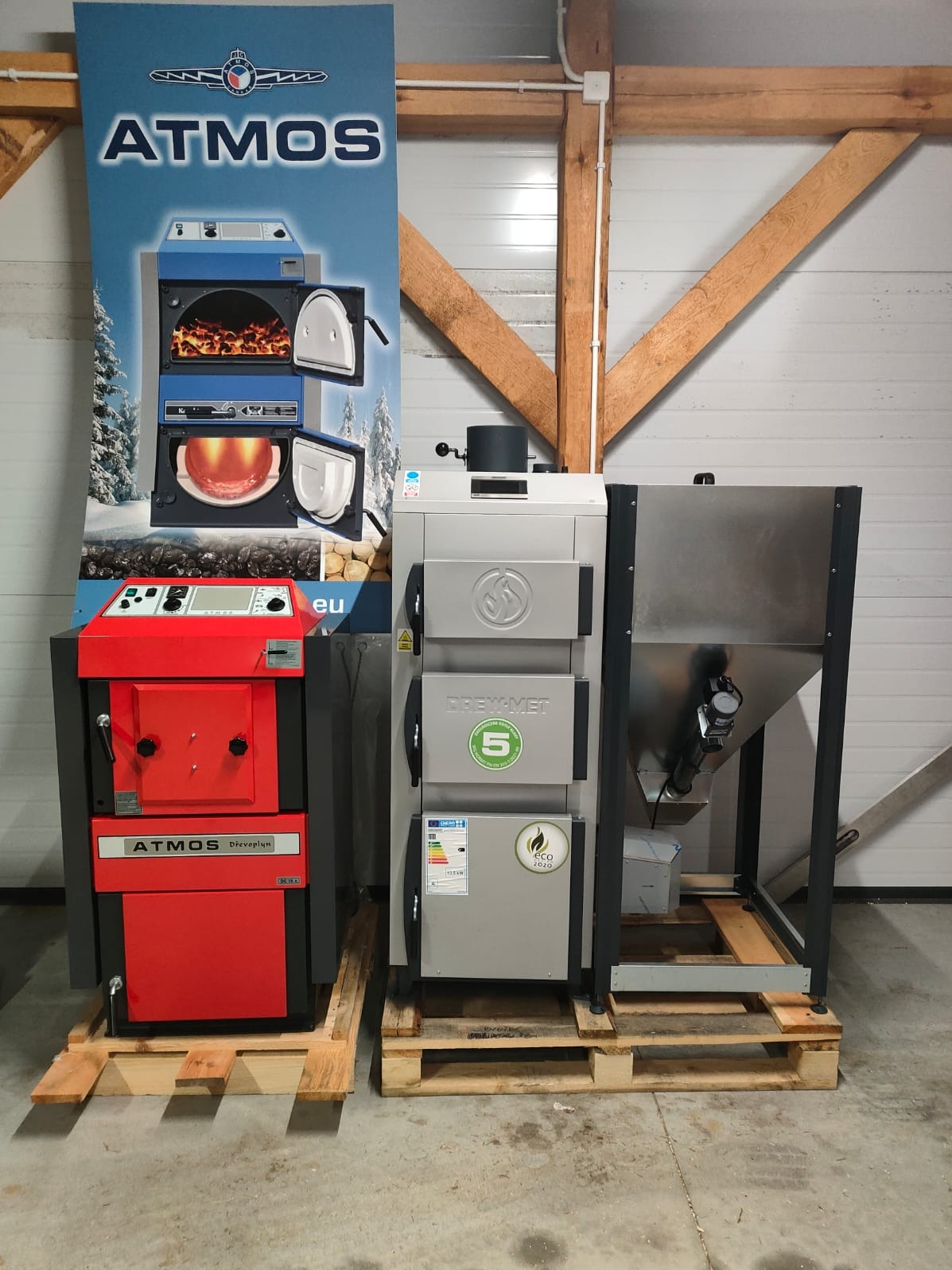 Dwa piece: czerwony Atmos na palecie obok baneru reklamowego Atmos, oraz szary Drew-Met z podajnikiem, oba stoją w hali.