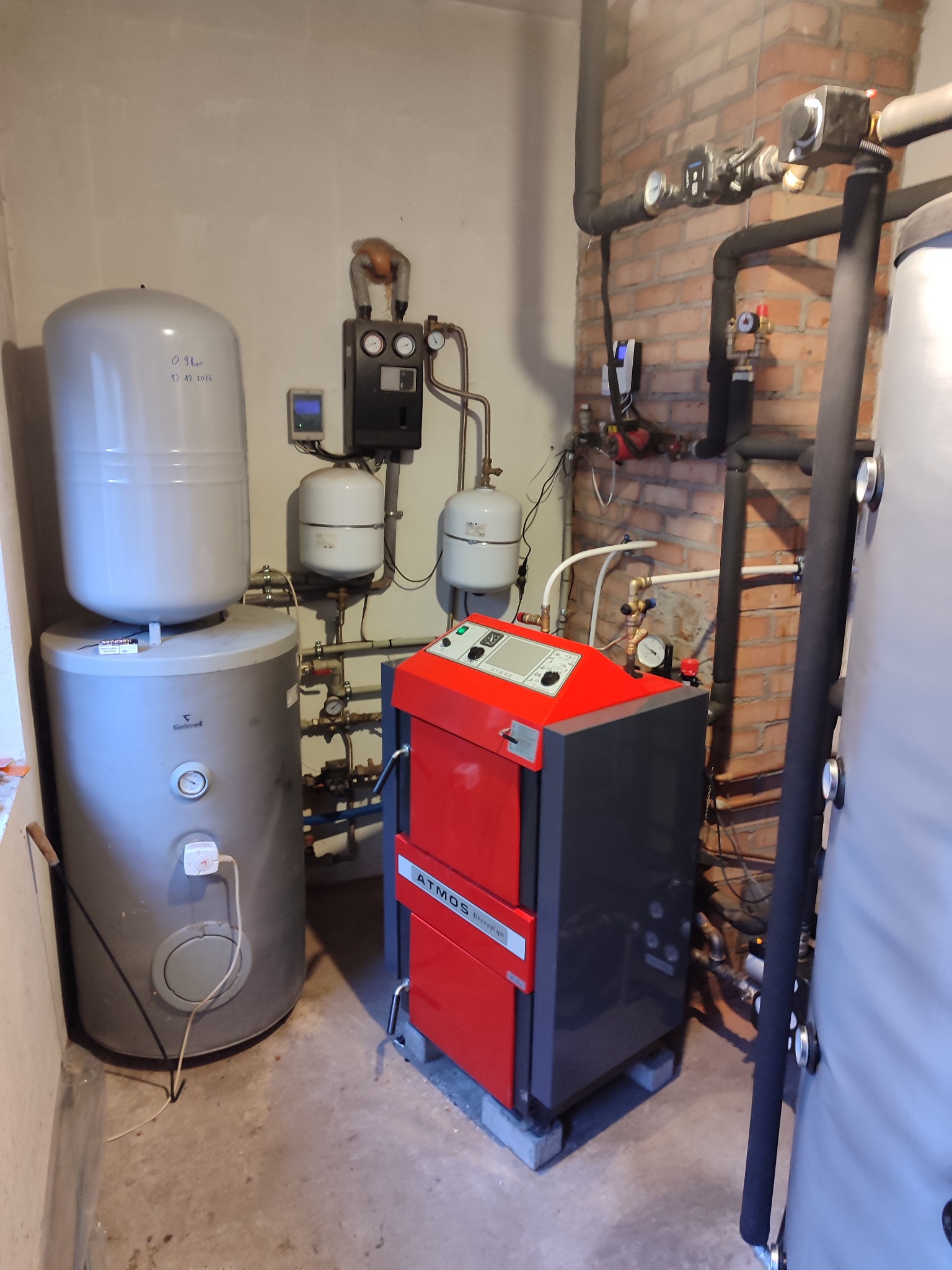 Kotłownia z piecem na paliwo stałe Atmos, zbiornikami buforowymi, instalacją hydrauliczną i sterownikami, widoczna ceglana ściana.