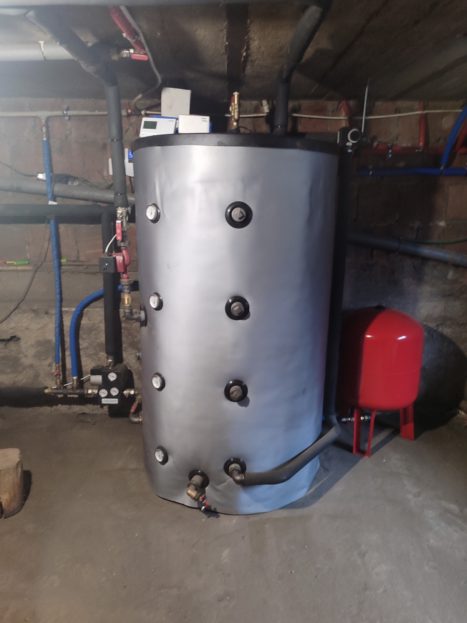 Szary akumulator ciepła z licznymi manometrami, zainstalowany w piwnicy z widocznymi rurami instalacji grzewczej i czerwonym zbiornikiem wyrównawczym.