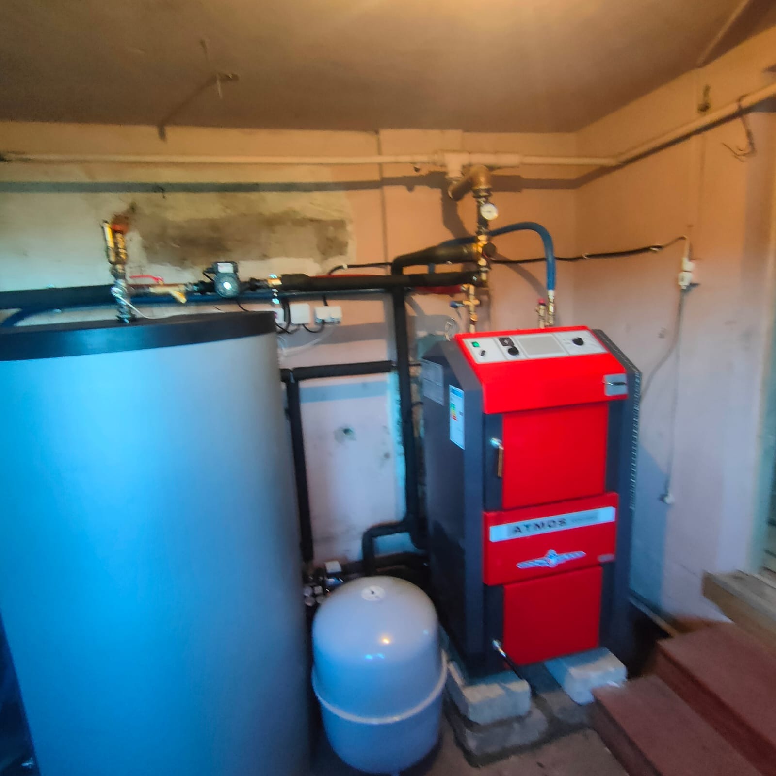 Czerwono-szary piec CO marki Atmos, zbiornik buforowy oraz naczynie przeponowe w kotłowni o ścianach z widocznymi śladami remontu.