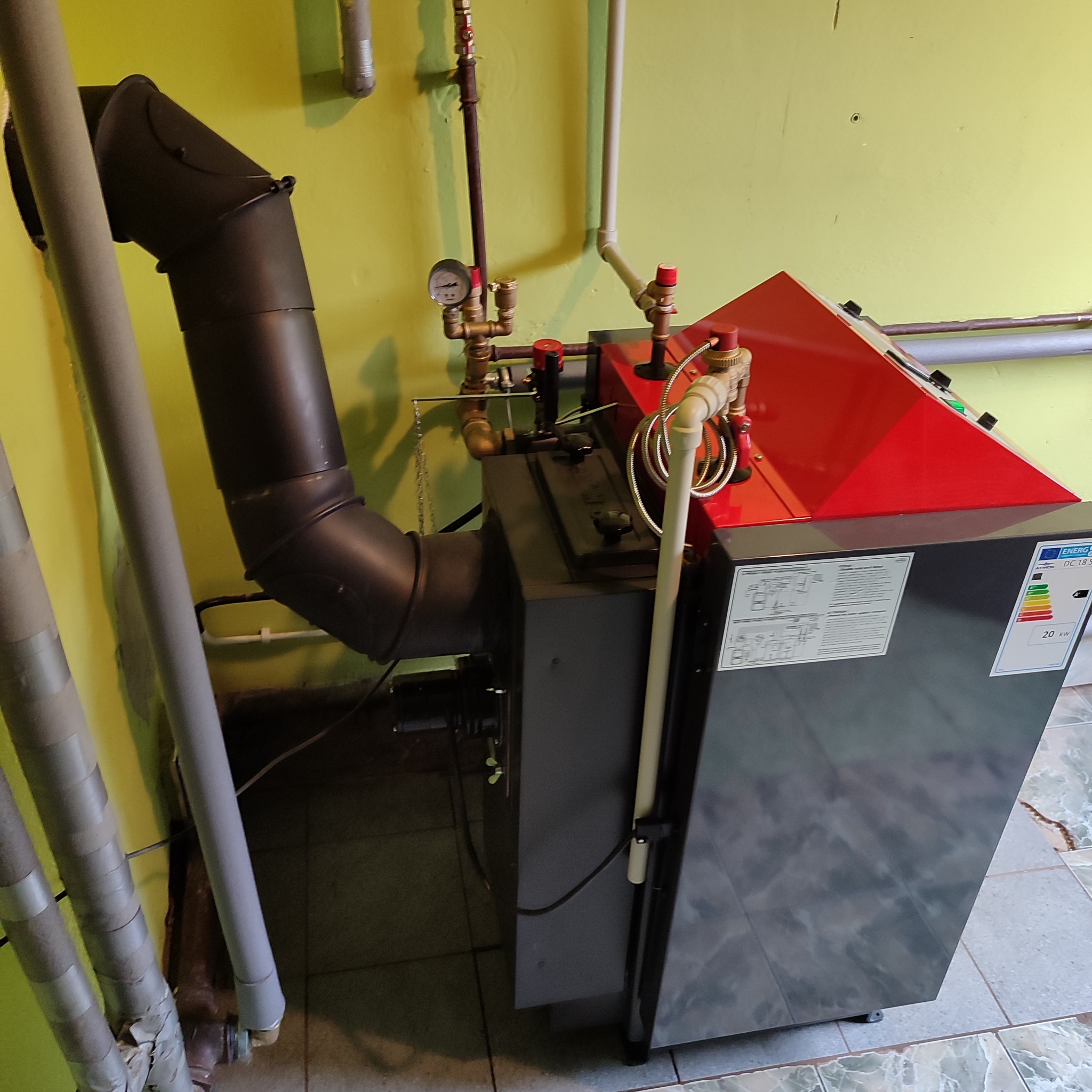 Nowoczesny piec CO z elementami w kolorze czerwonym, widoczny czarny komin odprowadzający spaliny, instalacja hydrauliczna i manometr. Na piecu naklejka z parametrami technicznymi.