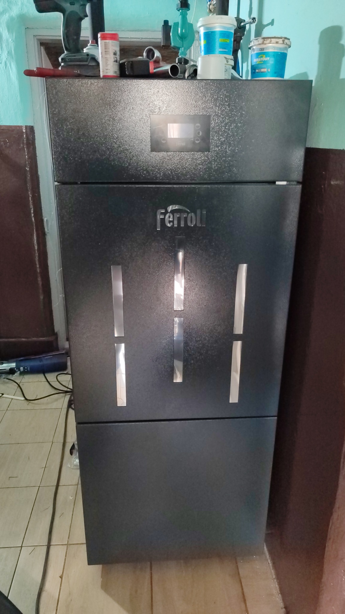 Ciemny piec Ferroli z wyświetlaczem i pionowymi, srebrnymi wstawkami, ustawiony na kafelkach, z narzędziami i pojemnikami na wierzchu, w pomieszczeniu o niebieskich ścianach.