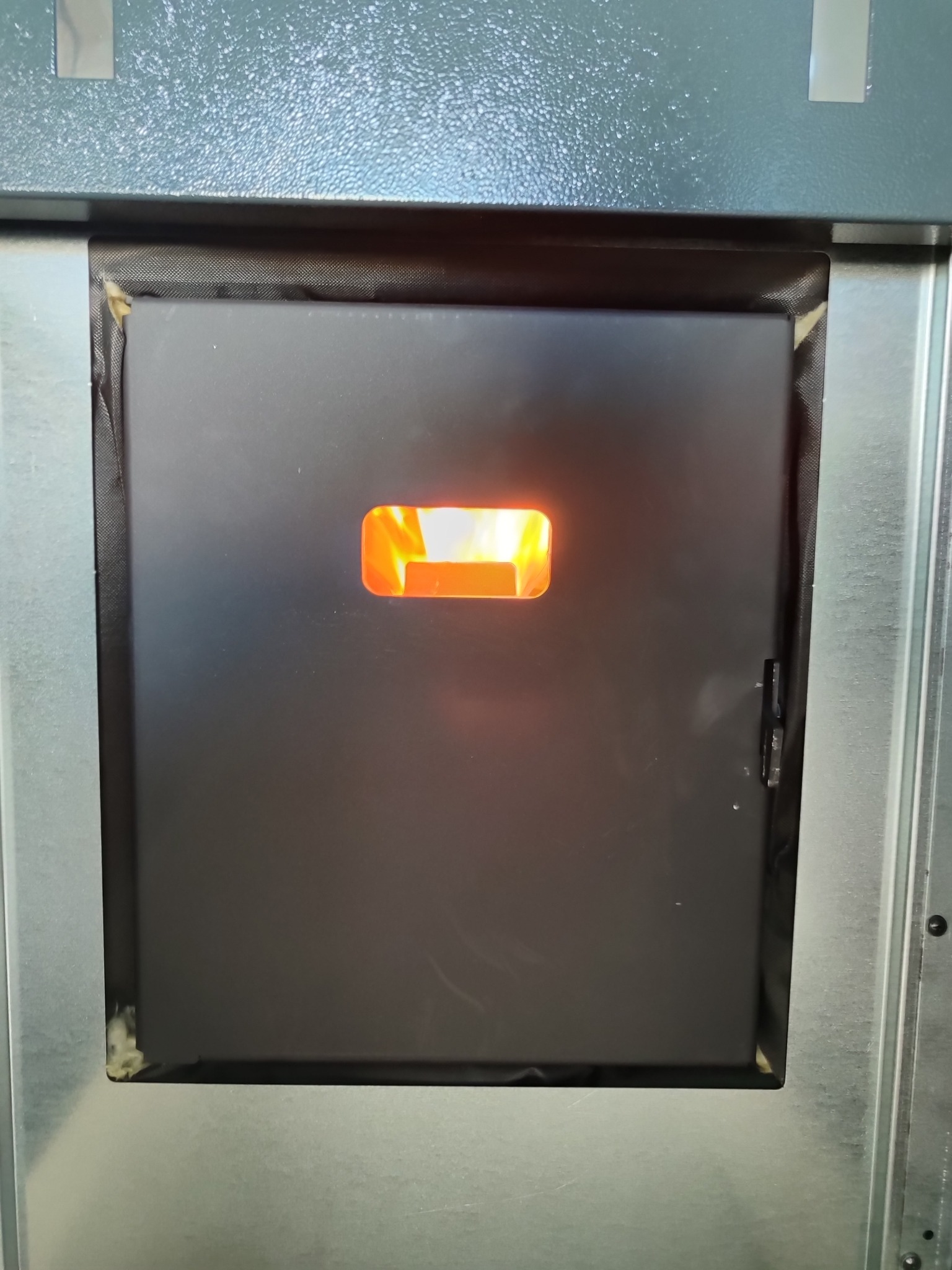 Zamknięte drzwiczki pieca z widocznym płomieniem przez otwór inspekcyjny, obudowa z szarej blachy, widoczne ślady uszczelnienia na krawędziach.