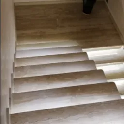 Schody z podświetleniem LED na każdym stopniu, wykonane z beżowego kamienia, z rośliną doniczkową na górnym podeście.