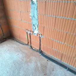 Ściana z czerwonej cegły z wyżłobieniami wypełnionymi zaprawą, w których umieszczono puszki elektryczne; kable w peszlach przymocowane do ściany i podłogi.