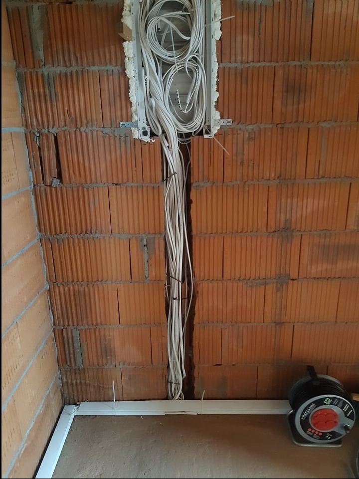 Wiązka przewodów elektrycznych wychodząca z puszki instalacyjnej w ścianie z czerwonej cegły, zabezpieczona opaskami zaciskowymi, z widocznym fragmentem podłogi i przedłużaczem bębnowym w prawym...