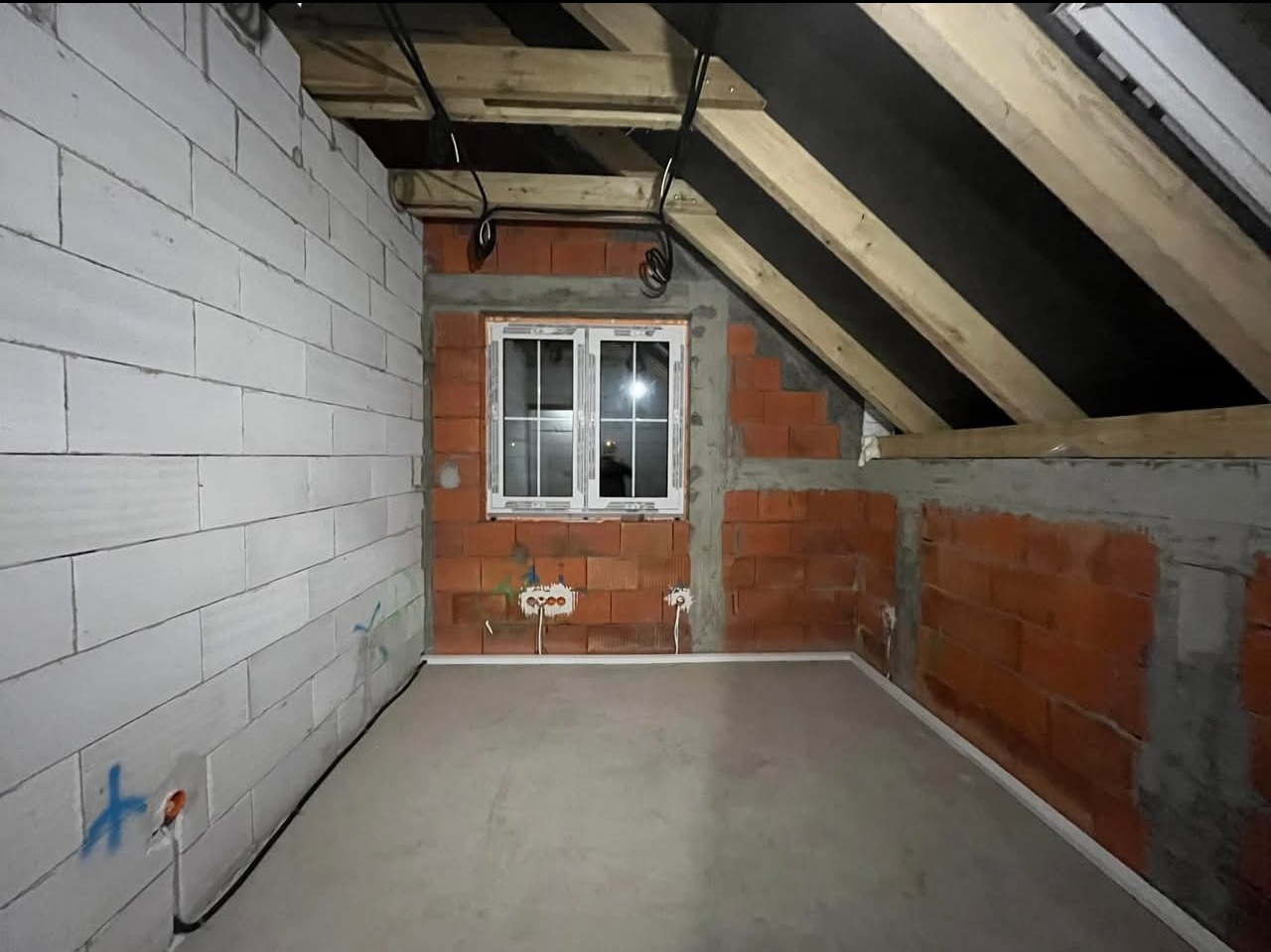 Surowy stan poddasza z widocznymi przewodami elektrycznymi pod sufitem, ścianami z czerwonej cegły i białego betonu komórkowego oraz oknem w białej ramie.