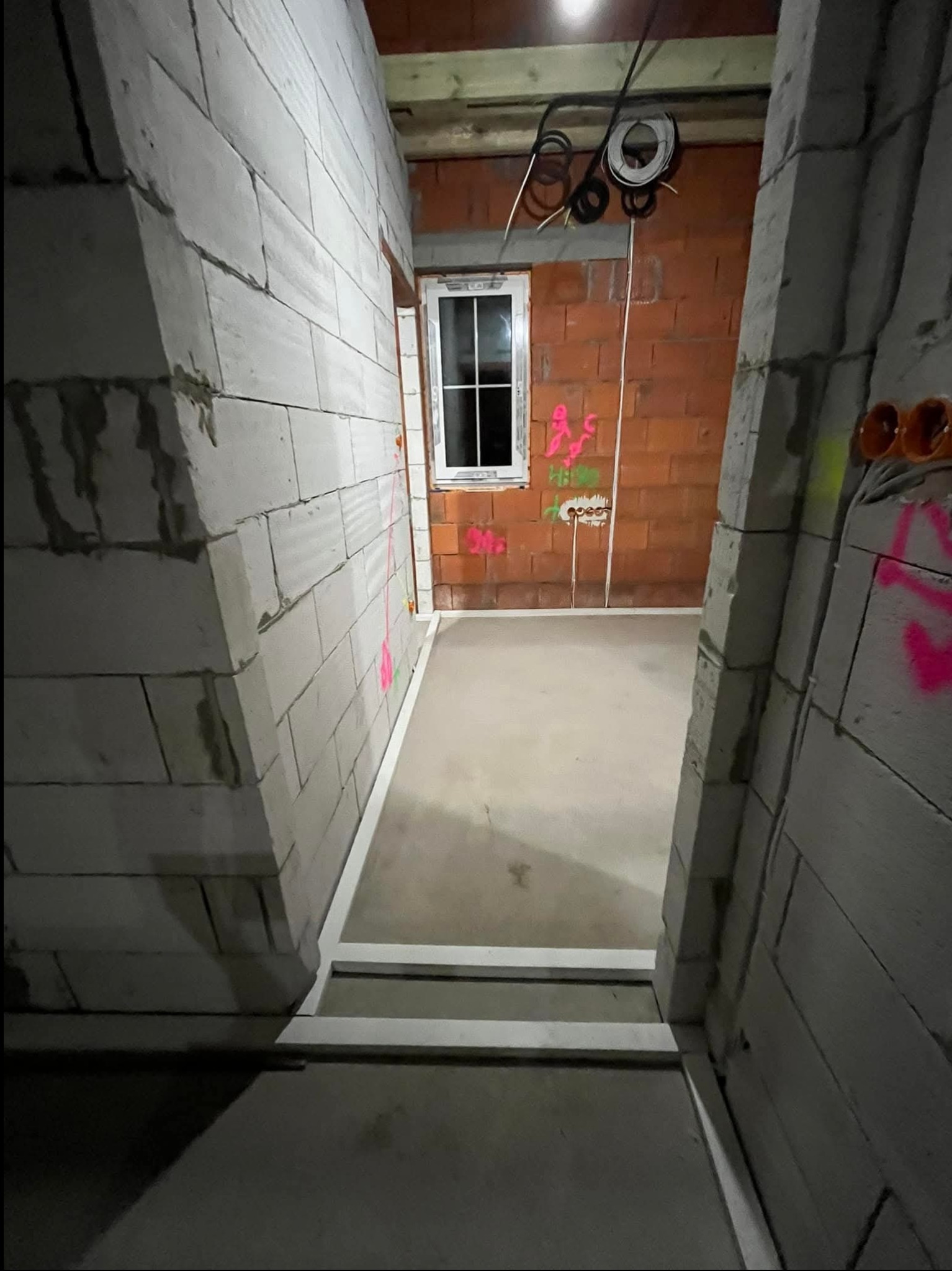 Surowy stan wnętrza w Namysłowie z widocznymi przewodami elektrycznymi zwisającymi z sufitu, instalacją podtynkową w ścianie z cegły oraz oznaczeniami wykonanymi różową farbą.