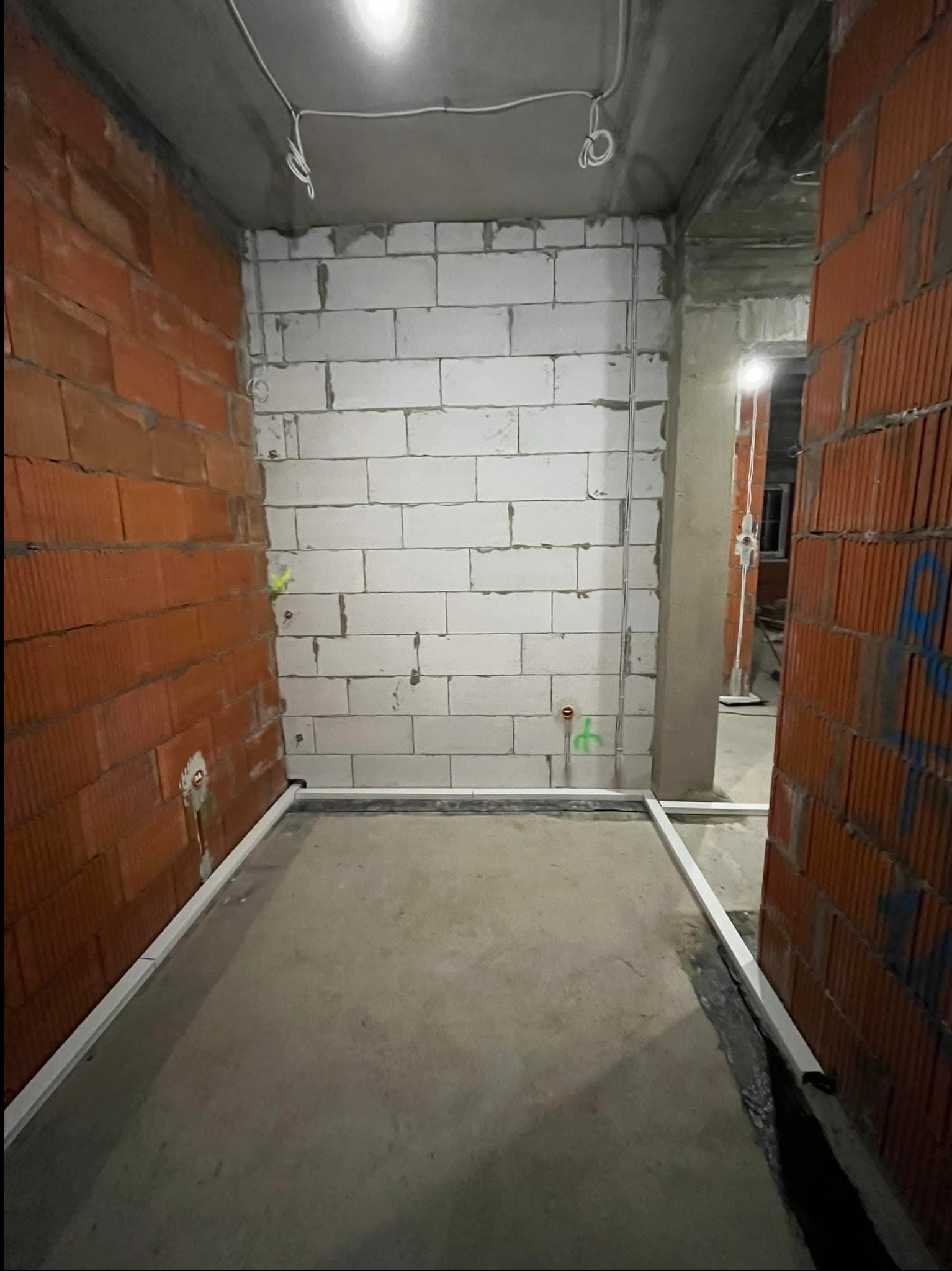 Wnętrze w trakcie prac wykończeniowych: ściany z czerwonej cegły i białego bloczka, widoczne przewody elektryczne wystające ze ściany, listwy przypodłogowe zamontowane wzdłuż ścian, surowy beton...