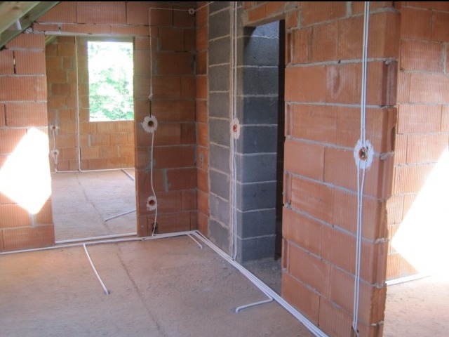 Surowy stan budynku w trakcie rozprowadzania instalacji elektrycznej: widoczne kable prowadzone po ścianach z czerwonej cegły i podłodze, puszki instalacyjne zamontowane w ścianach, okno w tle...