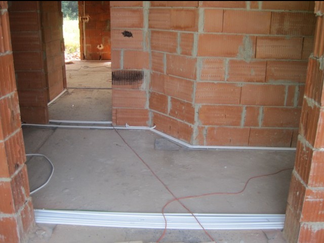 Wnętrze surowego budynku w trakcie prac, widoczne kable elektryczne prowadzone wzdłuż podłogi i ścian, zabezpieczone listwami przypodłogowymi, ściany z czerwonej cegły.