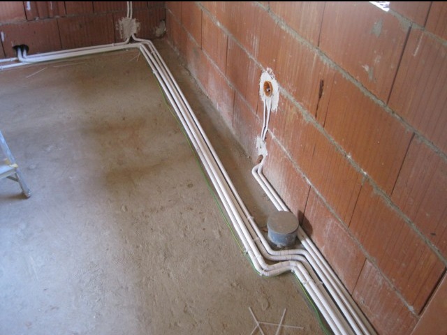 Białe rury instalacji elektrycznej ułożone wzdłuż ceglanej ściany w surowym stanie, widoczna puszka instalacyjna i fragment drabiny.