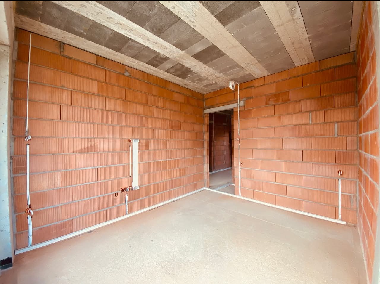 Surowy stan pokoju z cegły z widocznymi przewodami elektrycznymi w peszlach, przygotowanymi do dalszych prac wykończeniowych, betonowa wylewka na podłodze i surowy strop z betonowych płyt.