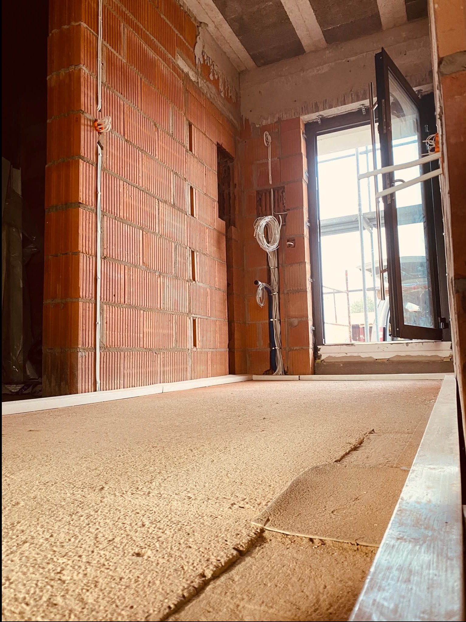 Wnętrze w trakcie budowy z widoczną ścianą z czerwonej cegły dziurawki, na której poprowadzono przewody elektryczne, oraz otwartym oknem z widokiem na rusztowanie.