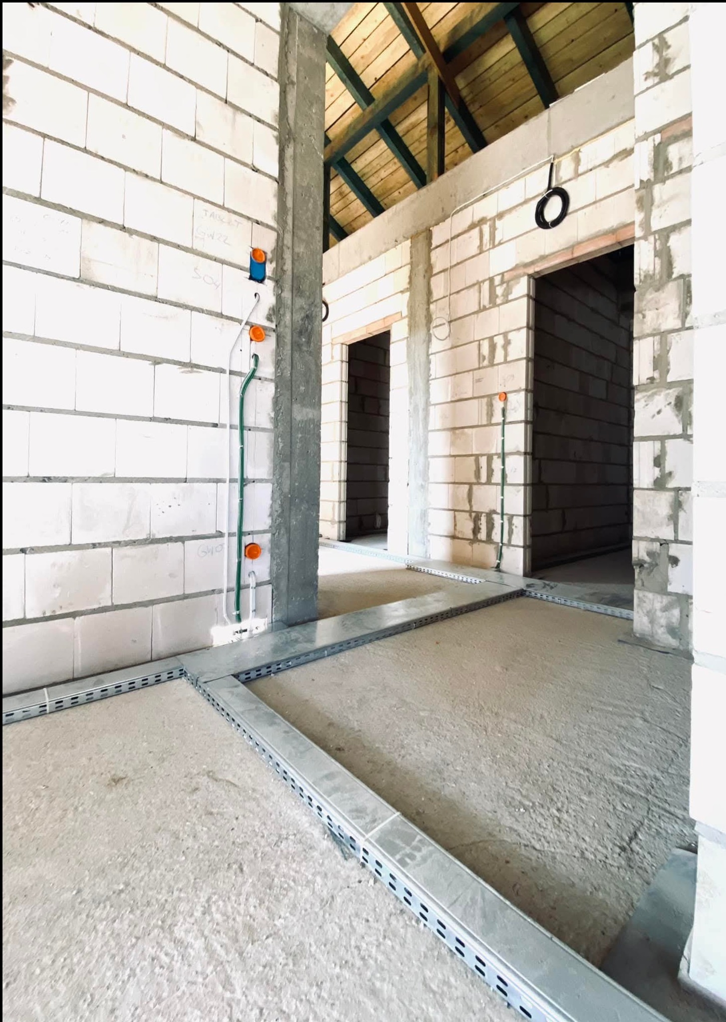 Surowy stan wnętrza budynku w trakcie prac elektrycznych: widoczne kable w peszlach, puszki elektryczne w ścianie z betonu komórkowego, trasy kablowe na podłodze, surowy strop drewniany.