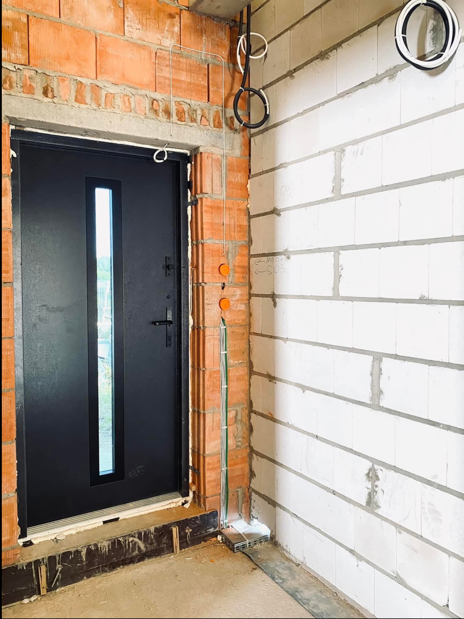 Czarne drzwi wejściowe z małym okienkiem, zamontowane w surowym murze z cegły, widoczne przewody elektryczne prowadzone po ścianie i nadprożu, ściana z białych bloczków z widocznymi oznaczeniami...