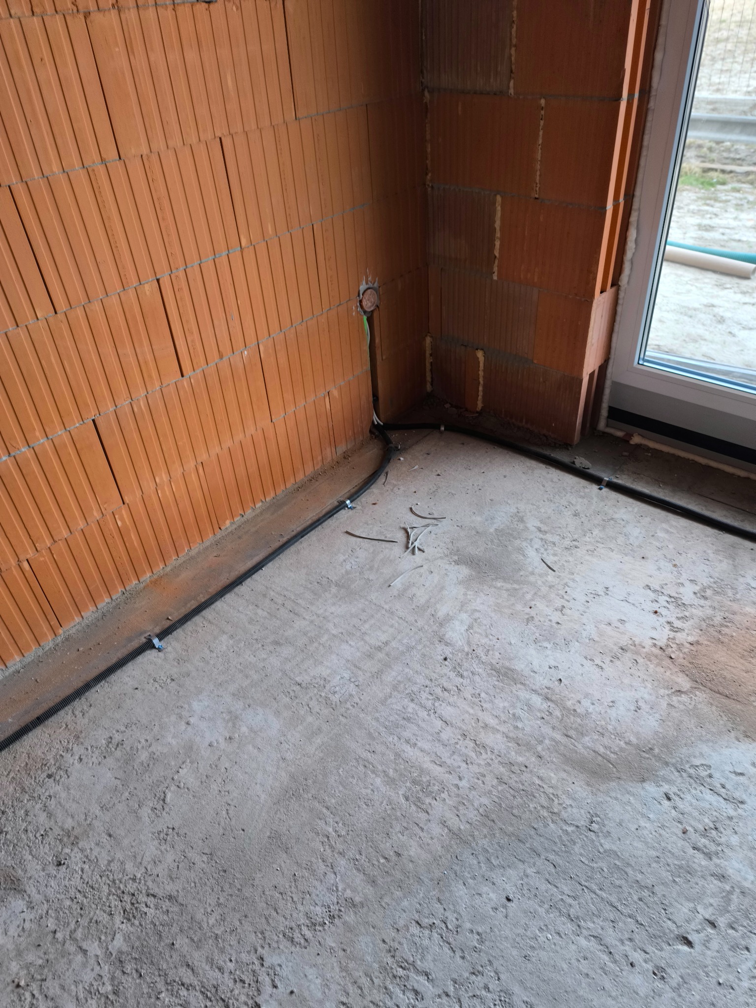 Kabel elektryczny w peszlu, przymocowany klamrami do ściany z cegły dziurawki, w surowym stanie, przed wylewką posadzki. Widoczne pozostałości po kablach i otwór w ścianie pod puszkę elektryczną.