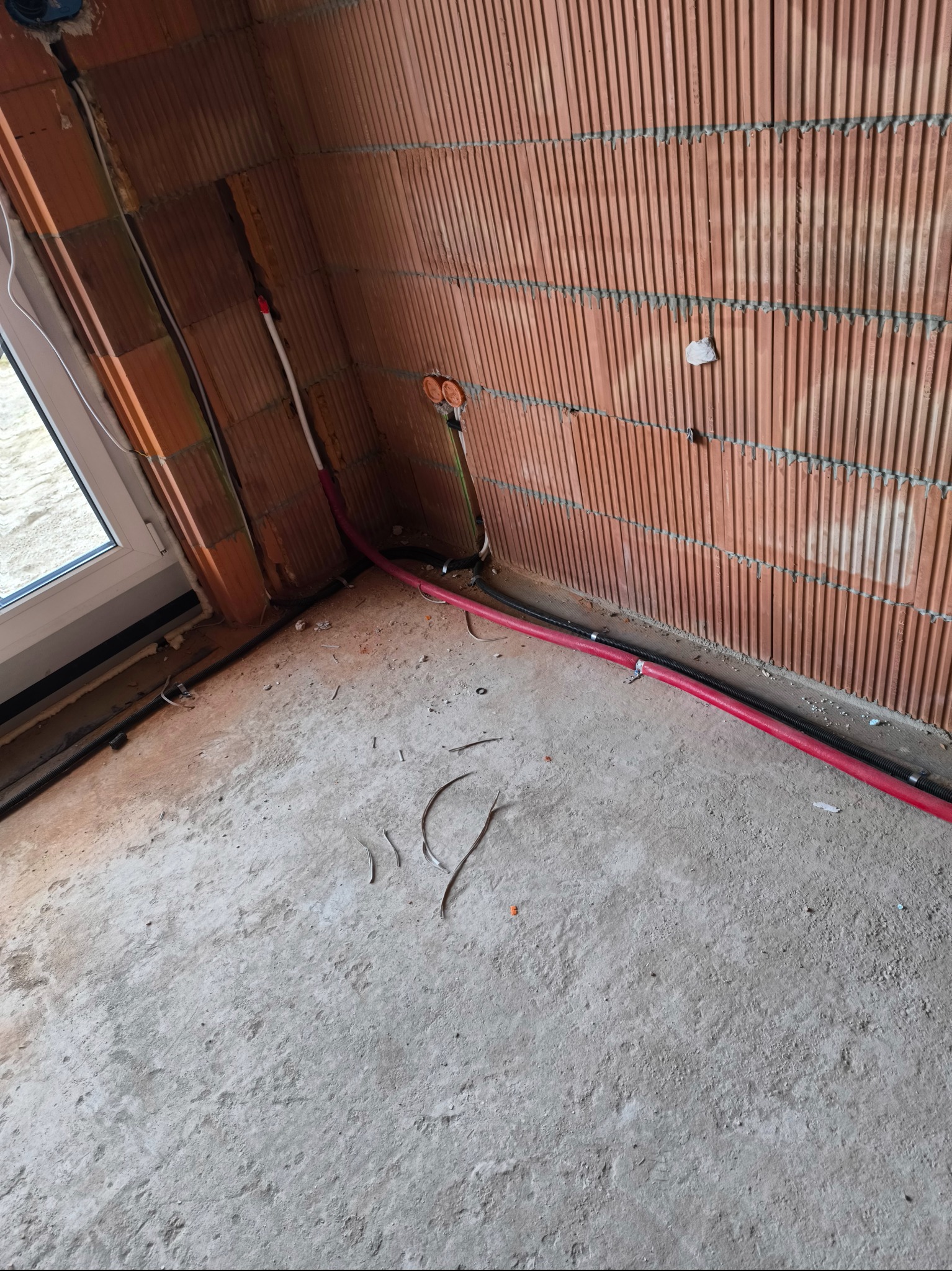 Wewnętrzne okablowanie elektryczne w nowym budynku z widocznymi przewodami w kolorze czerwonym i czarnym prowadzonymi wzdłuż ściany z pustaków ceramicznych.