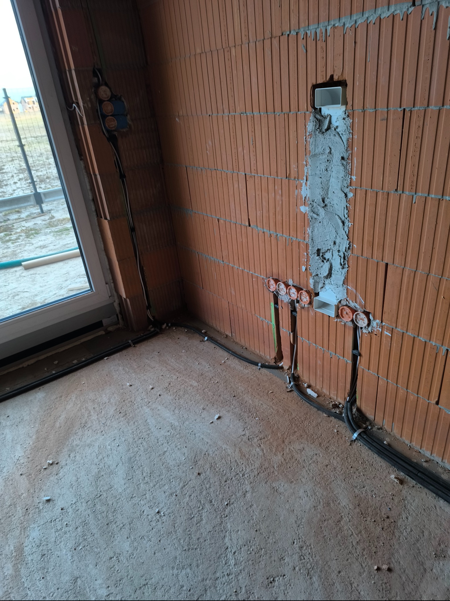 Wewnętrzne okablowanie elektryczne w surowym budynku z czerwonej cegły, widoczne puszki instalacyjne, przewody prowadzone wzdłuż ściany i pod podłogą, oraz zamurowana rura instalacyjna.