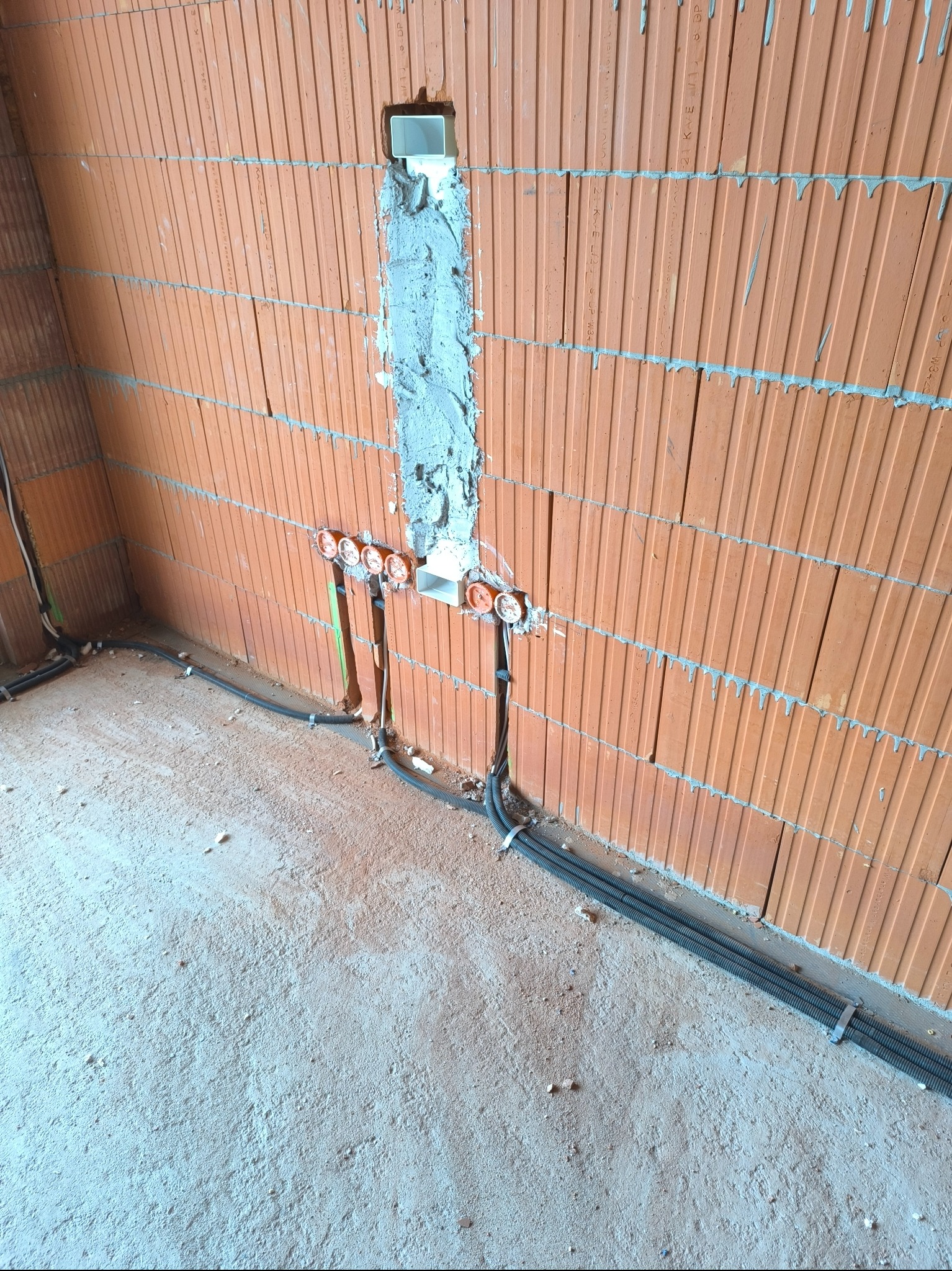 Ściana z czerwonej cegły z wyżłobieniami wypełnionymi zaprawą, w których umieszczono puszki elektryczne; kable w peszlach przymocowane do ściany i podłogi.