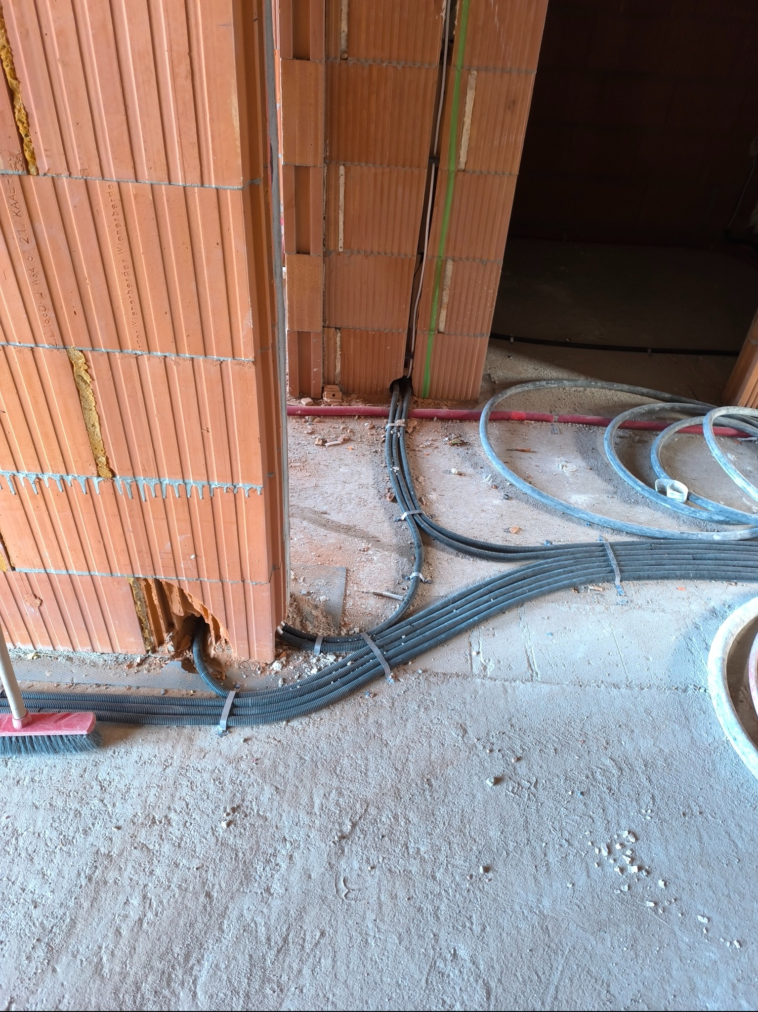 Wiązka przewodów elektrycznych w peszlach wychodząca z otworu w murze z cegły ceramicznej, zabezpieczona opaskami zaciskowymi, widoczne elementy instalacji podłogowej i ściany w surowym stanie.