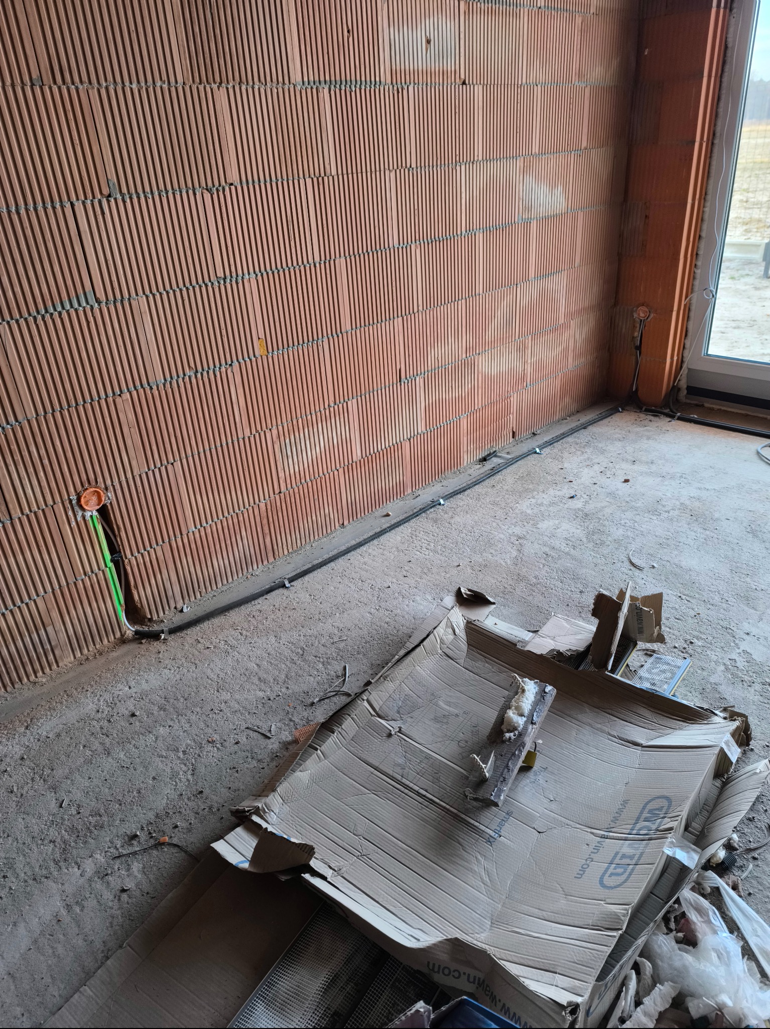 Wnętrze w trakcie prac wykończeniowych: przewody elektryczne w peszlu, prowadzone wzdłuż ściany z cegły ceramicznej dziurawki, widoczne puszki instalacyjne i resztki materiałów budowlanych...