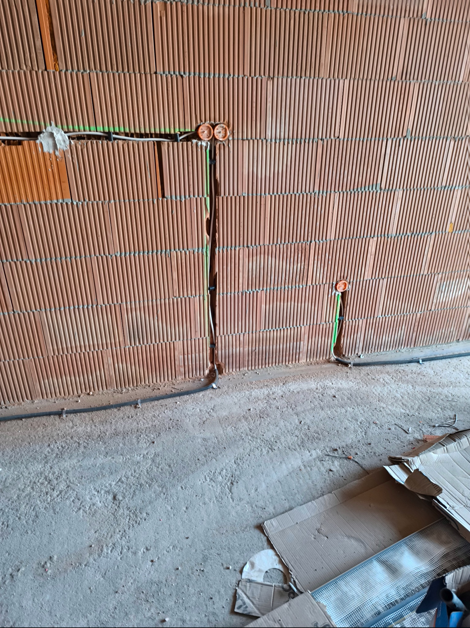 Ściana z pustaków ceramicznych z bruzdami pod przewody elektryczne, widoczne puszki instalacyjne, kable w peszlach i fragment podłogi w stanie surowym.