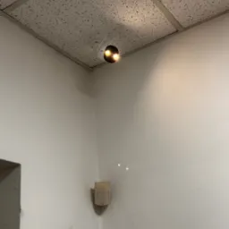 Kamera monitoringu zamontowana pod sufitem podwieszanym w rogu pomieszczenia, obok czujnika ruchu na ścianie.