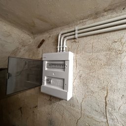MODERON SYSTEM Michał Tkocz - Biała skrzynka rozdzielcza natynkowa z otwartymi drzwiczkami, zamontowana na szarej, popękanej ścianie w piwnicy. Nad skrzynką widoczne trzy szare rury prowadzone wzdłuż sufitu, przymocowane...