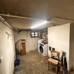 MODERON SYSTEM Michał Tkocz - Natynkowe rury i puszki elektryczne prowadzone po suficie i ścianach piwnicy, widoczna lampa jarzeniowa, pralka oraz prowizoryczne miejsce pracy z krzesłem i narzędziami.
