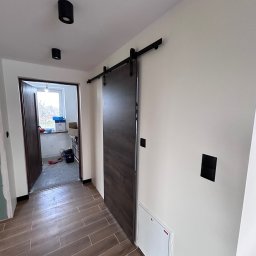MODERON SYSTEM Michał Tkocz - Wnętrze korytarza z przesuwnymi drzwiami w stylu stodoły, brązowe płytki podłogowe imitujące drewno, czarne lampy sufitowe i włączniki, widoczna skrzynka rewizyjna.