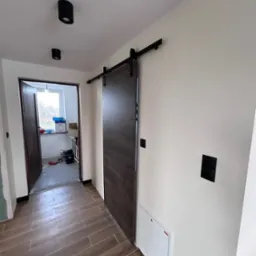 Wnętrze korytarza z przesuwnymi drzwiami w stylu stodoły, brązowe płytki podłogowe imitujące drewno, czarne lampy sufitowe i włączniki, widoczna skrzynka rewizyjna.