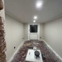 MODERON SYSTEM Michał Tkocz - Pokój po remoncie: świeżo pomalowane ściany w odcieniach szarości, widoczne puszki z farbą na zabezpieczonej podłodze, fragment odsłoniętej cegły na ścianie po lewej stronie, widoczne gniazdka...