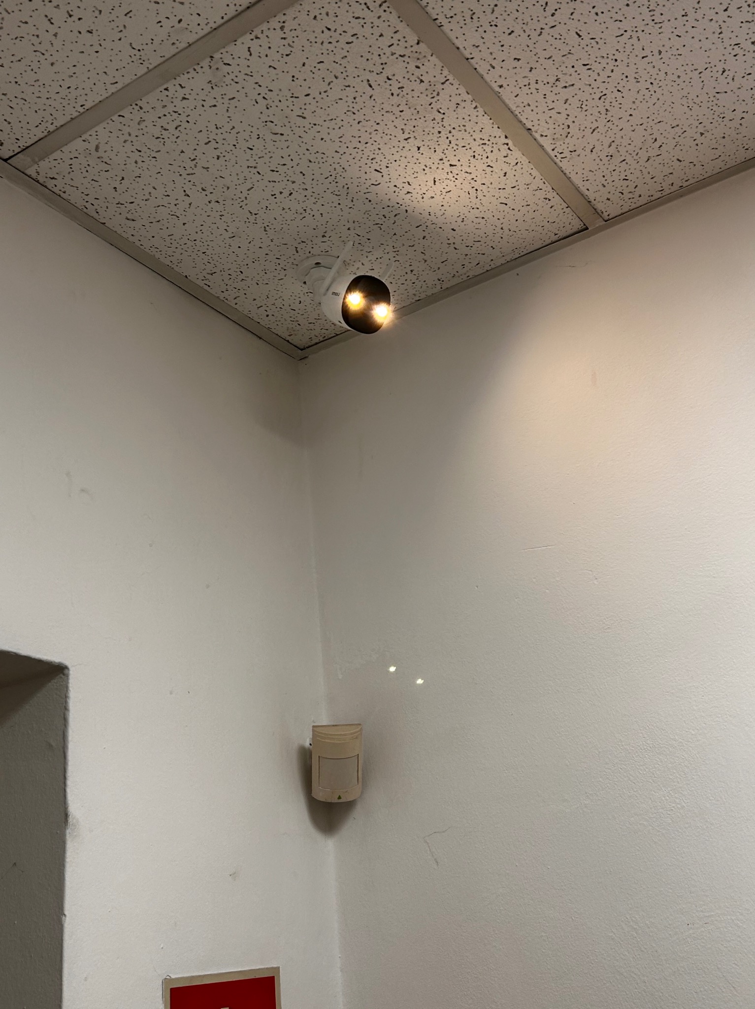 Kamera monitoringu zamontowana pod sufitem podwieszanym w rogu pomieszczenia, obok czujnika ruchu na ścianie.