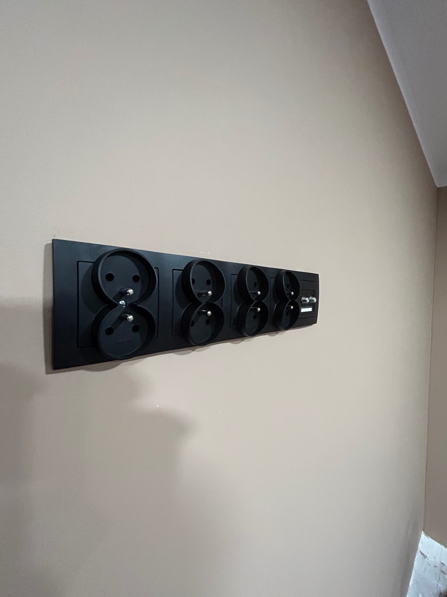 Czarny panel z czterema podwójnymi gniazdami elektrycznymi i dwoma portami USB, zamontowany na beżowej ścianie.