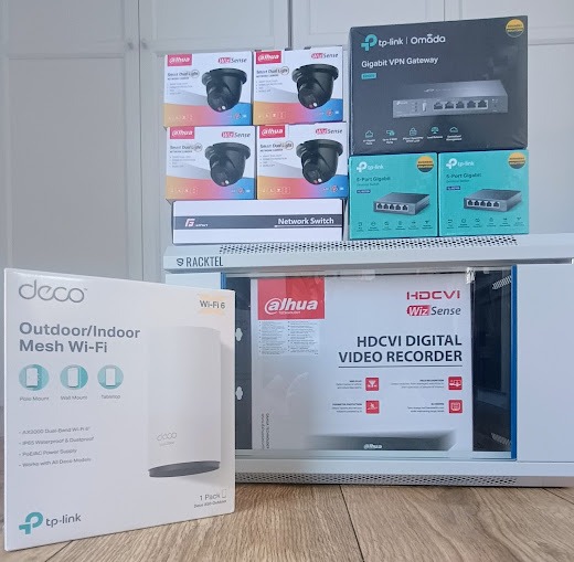 Pudełka z kamerami, rejestratorem wideo, routerem VPN, switchem sieciowym i punktem dostępowym Wi-Fi, ułożone na półce, gotowe do instalacji.