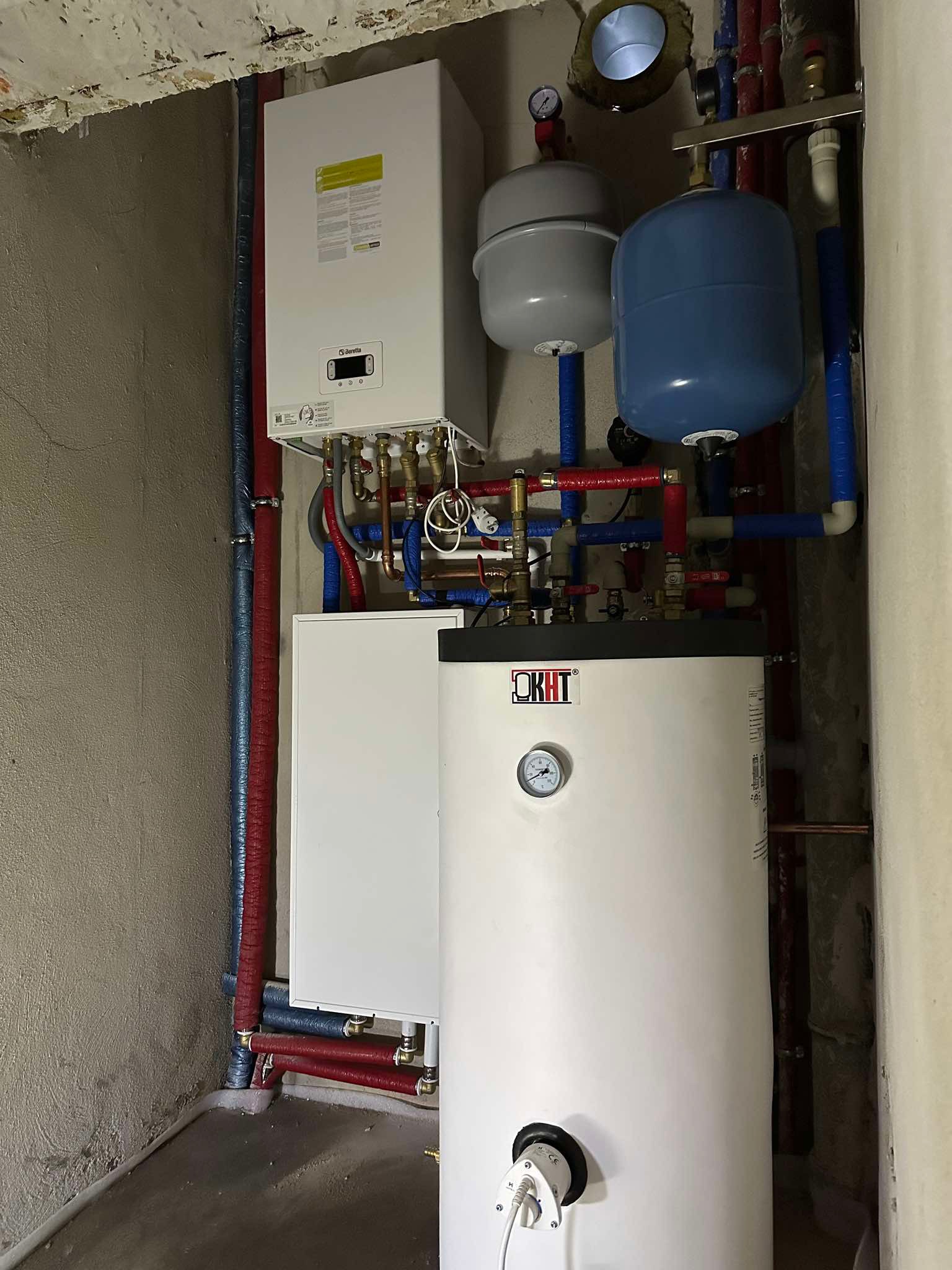 Zestaw urządzeń grzewczych w piwnicy: biały piec gazowy Beretta, zbiornik OKHT z manometrem, dwa zbiorniki wyrównawcze, orurowanie w izolacji czerwonej i niebieskiej.