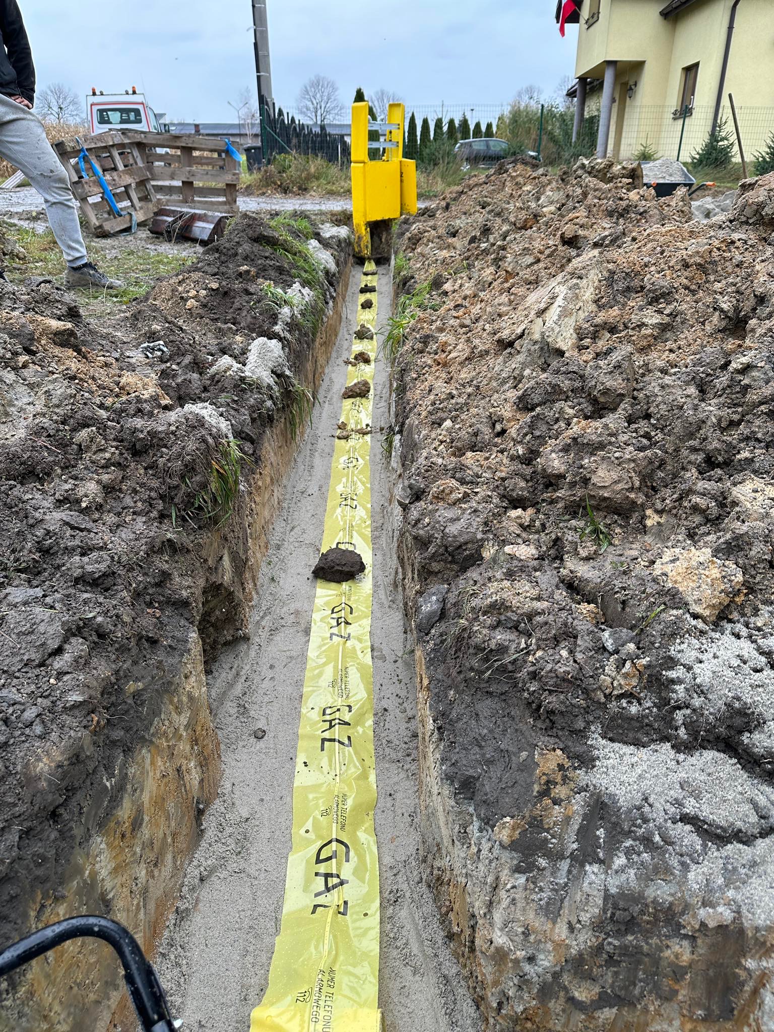 Wykop pod instalację gazową zabezpieczony żółtą folią ostrzegawczą z napisem 'GAZ', widoczna maszyna układająca rury.