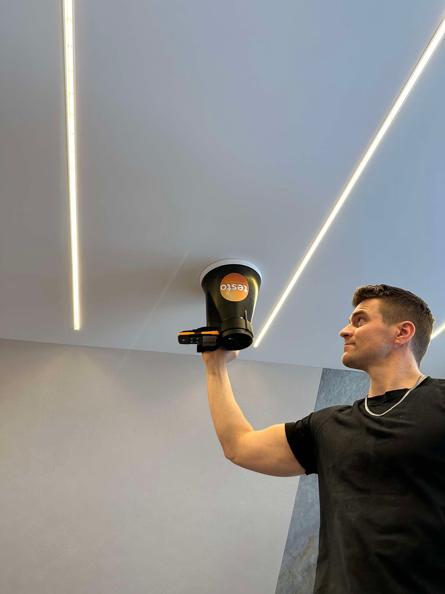 Mężczyzna mierzy przepływ powietrza anemometrem marki Testo przy suficie z oświetleniem liniowym LED.