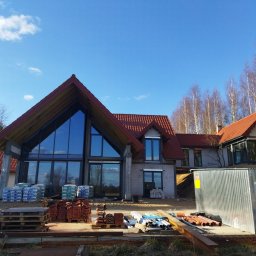 BUDMAR INVEST - Budowa domu w stanie surowym otwartym z widocznymi materiałami budowlanymi, czerwona dachówka, duże okna z refleksami nieba. W tle drzewa i błękitne niebo.