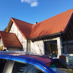 BUDMAR INVEST - Dom w trakcie budowy z czerwoną dachówką, widoczny fragment niebieskiego samochodu na pierwszym planie, worki z materiałami budowlanymi obok budynku.