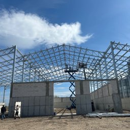 BUDMAR INVEST - Stalowa konstrukcja budynku w trakcie budowy z widocznymi fragmentami ścian z betonu komórkowego, podnośnikiem koszowym i pracownikami na zewnątrz.