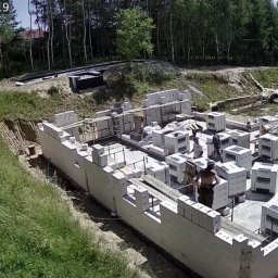 BUDMAR INVEST - Budowa fundamentów domu z bloczków betonowych na działce z widokiem na jezioro, w tle las. Widoczni pracownicy budowlani w trakcie pracy.