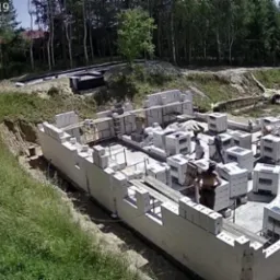 Budowa fundamentów domu z bloczków betonowych na działce z widokiem na jezioro, w tle las. Widoczni pracownicy budowlani w trakcie pracy.