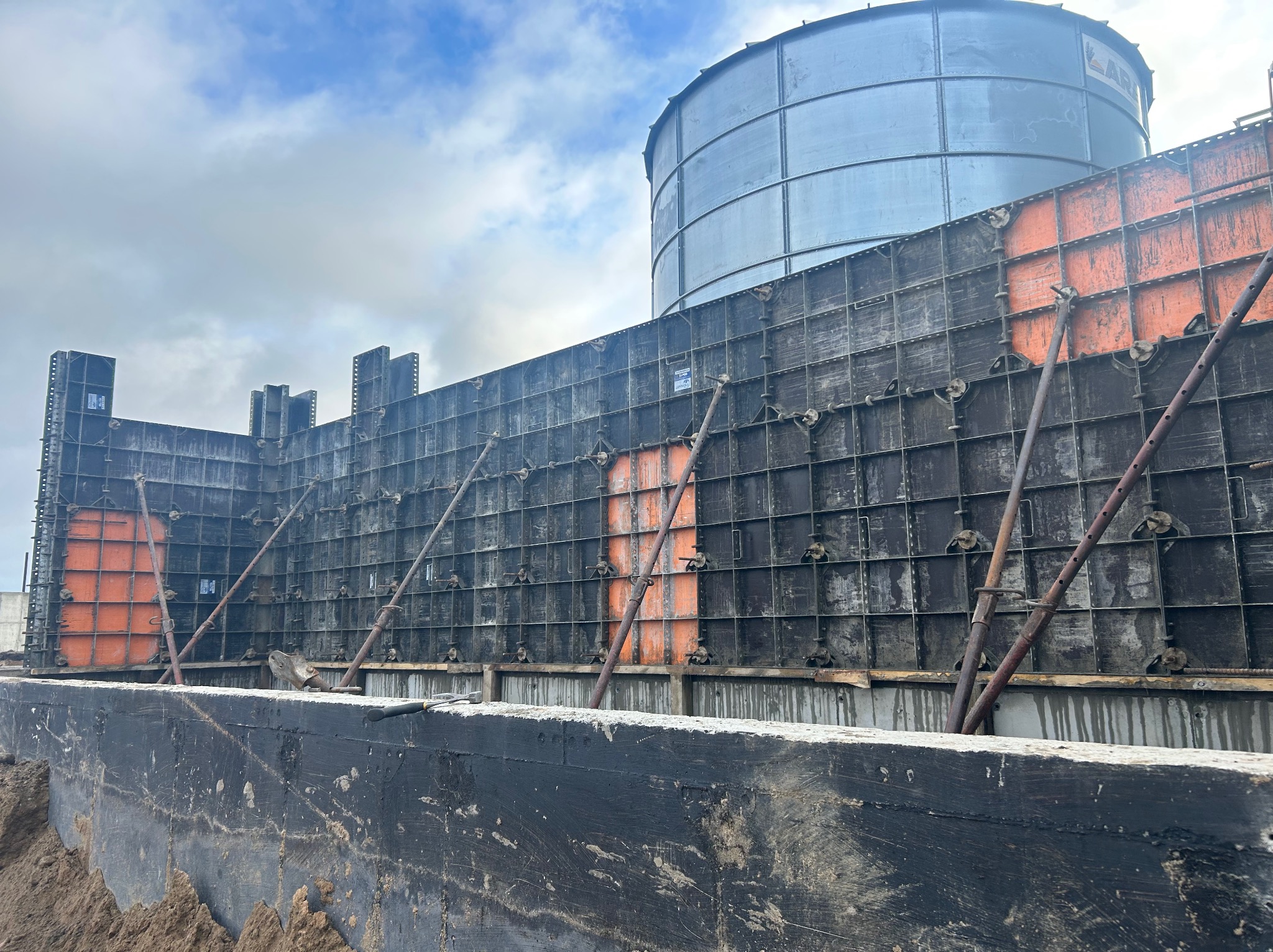 Szczegółowe ujęcie szalunku fundamentów z widocznym zbrojeniem, podpieranego skośnymi stemplami, na tle stalowego zbiornika i pochmurnego nieba.